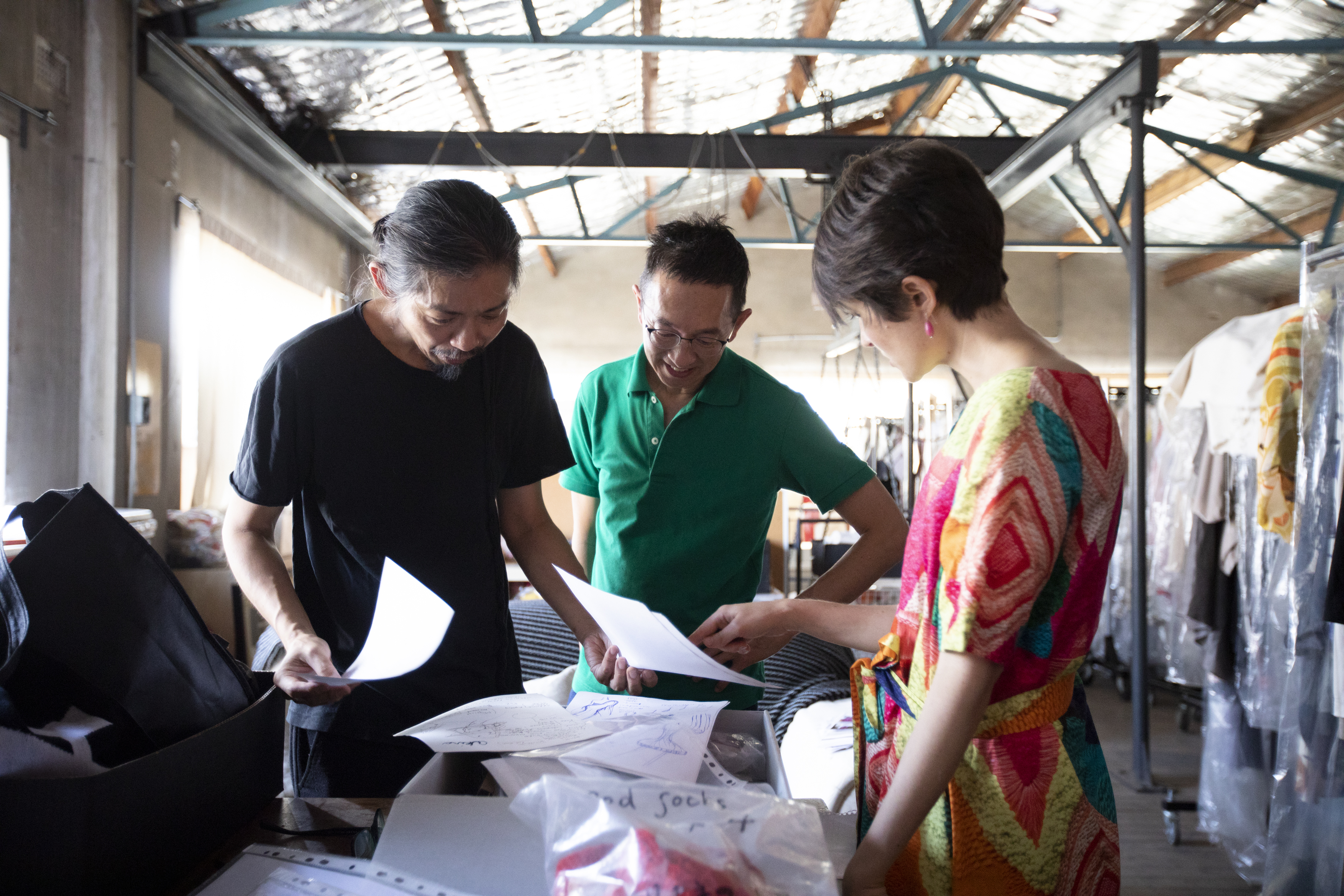 Two men and a woman stand around a table discussing fashion drawings and fabric samples. They are in the designer’s studio with garments hanging on racks in the background.